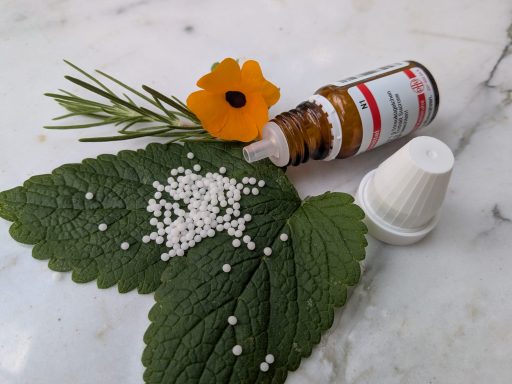 Braune Flasche mit Homöopathie-Kügelchen, grünen Blättern und einer orangefarbenen Blume.